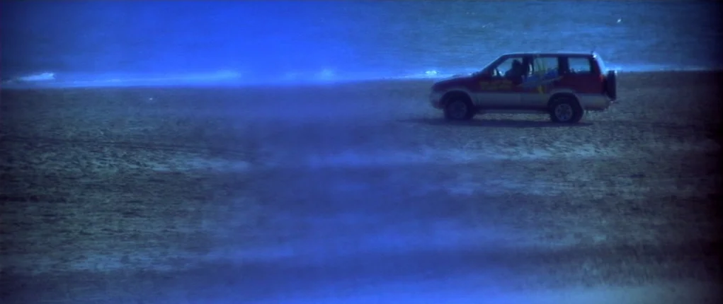 I Want You (1998) movie still: wide — A dark, blue-toned beach scene shows a vehicle parked near the tide line. Low, hazy light illuminates the sand…
