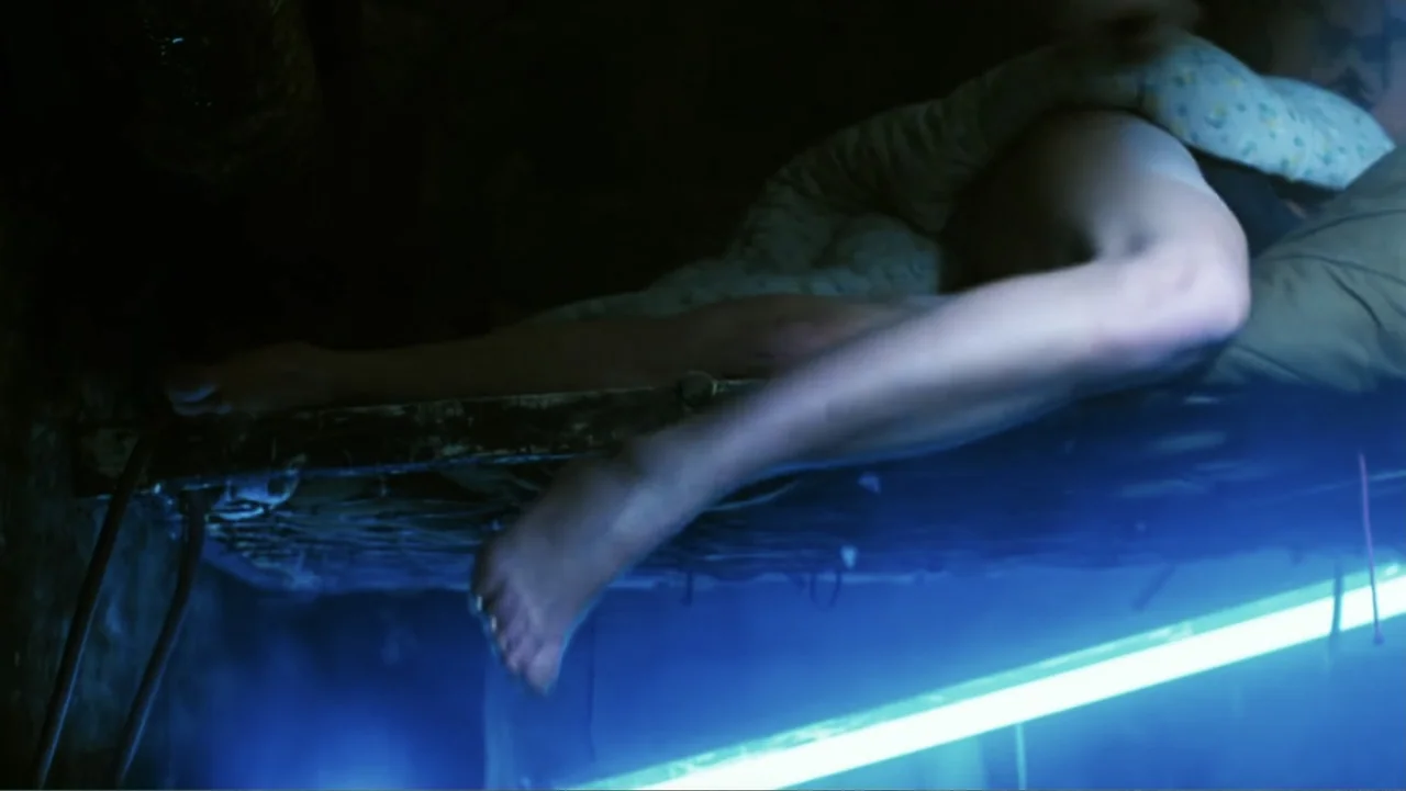 O-Bi, O-Ba: The End of Civilization (1985) movie still: medium — A dark, cramped space features a thin metal cot with a light-colored pillow. An…