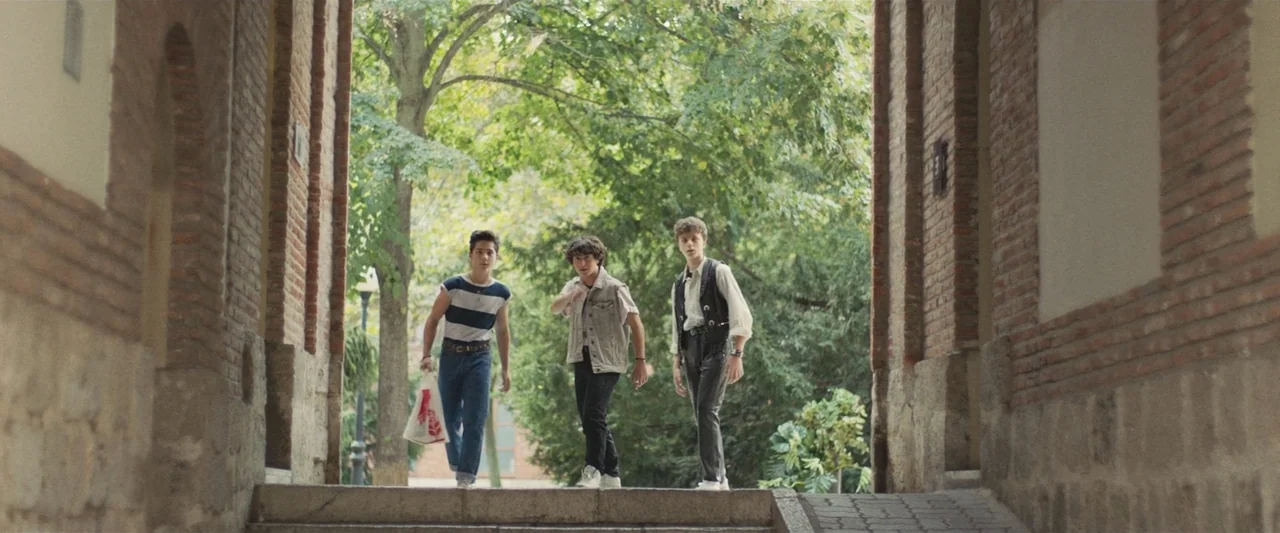 Voy a pasármelo bien (2022) movie still: fullbody — A stone tunnel leads into a lush park with green trees. Three teenagers walk side by side…