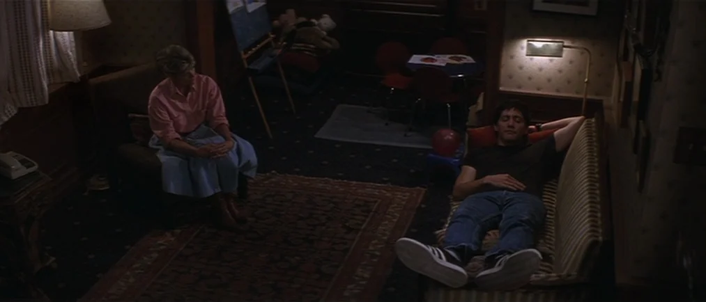 Donnie Darko movie still: wide — A dimly lit room contains patterned wallpaper and dark carpeting covering the floor. A woman sits in an armchair as…