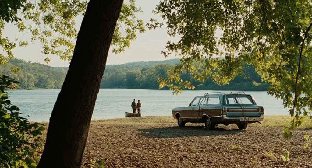 A Serious Man (2009) movie still: wide — A rural lakeshore features a parked vintage station wagon on a dirt bank. Two figures stand near a small…