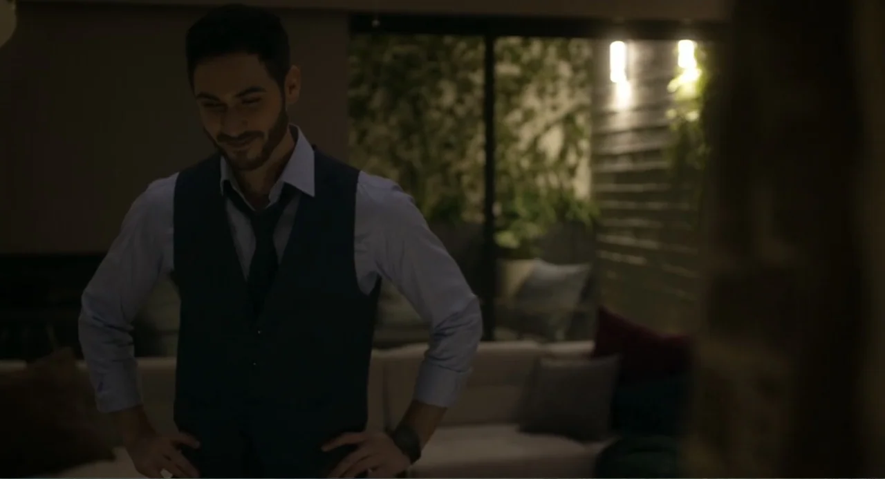 Straight (2024) movie still: medium — A dimly lit interior space with a couch and blurred background foliage visible through windows. A man in a…