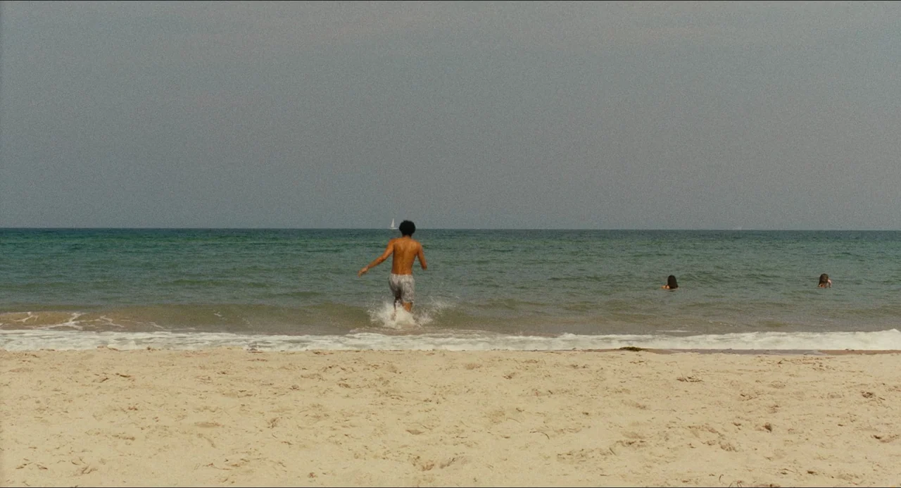 Afire (2023) movie still: wide — A wide sandy beach meets a calm, greenish-blue ocean under a grey overcast sky. A shirtless man runs into the…