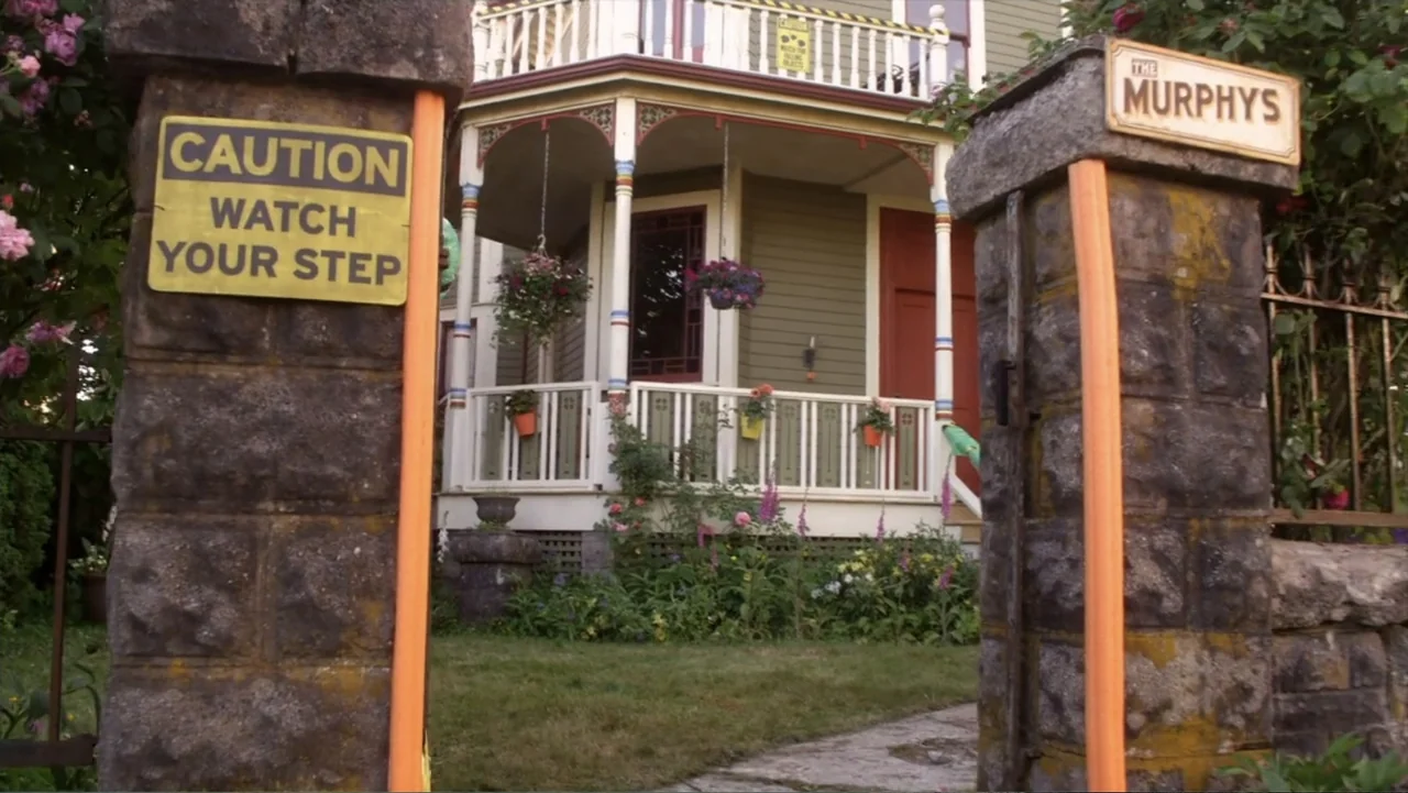 Jinxed (2013) movie still: establishing — A green residential house with a front porch sits behind two stone gate pillars. A sign on the pillar…