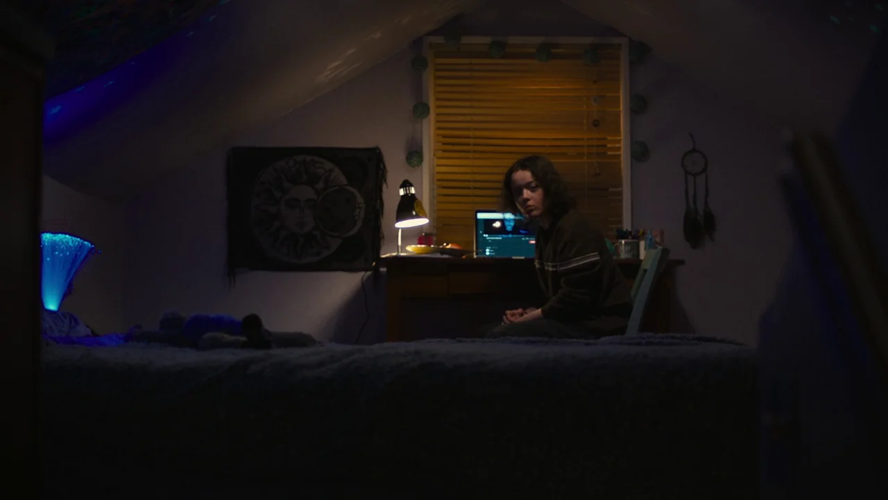 We’re All Going to the World’s Fair (2021) movie still: medium — A small bedroom in an attic space with sloped ceilings and dim purple ambient…