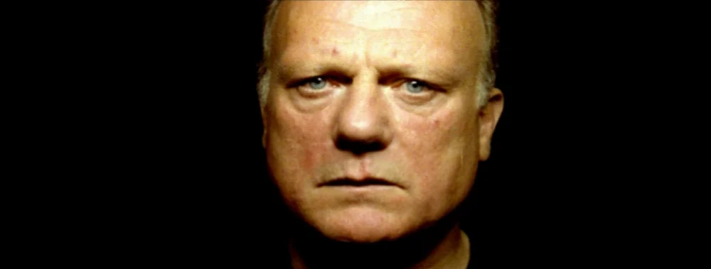 Carne (1991) movie still: closeup — A black, featureless background surrounds an older Caucasian man. The subject faces the camera directly with a…