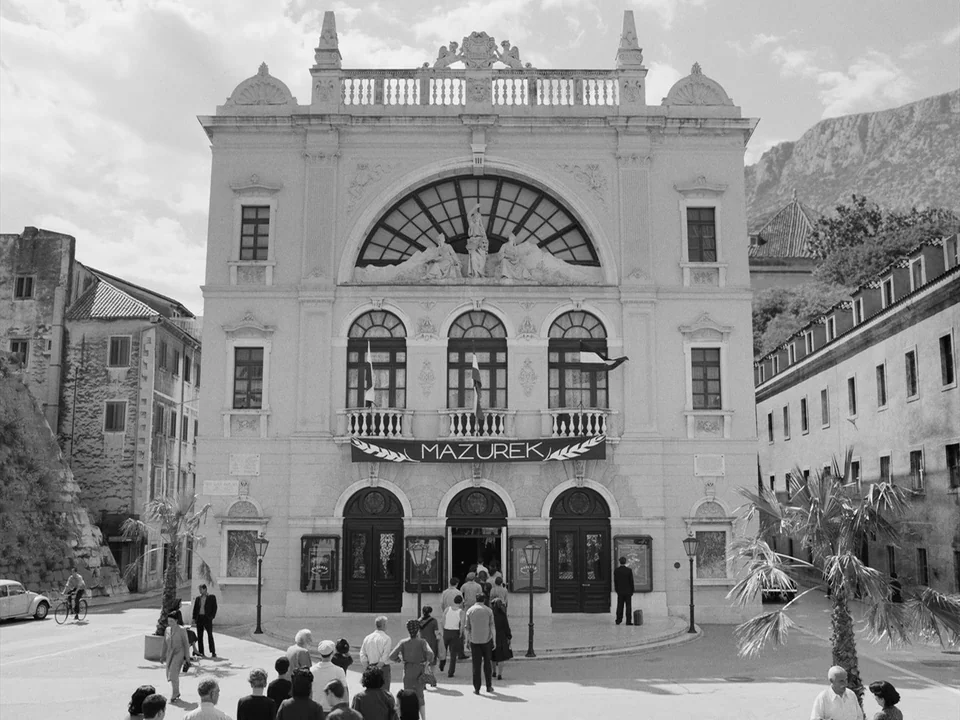 Cold War (2018) movie still: establishing — A historic stone theater with a grand arched window stands in a public plaza. People walk toward the…