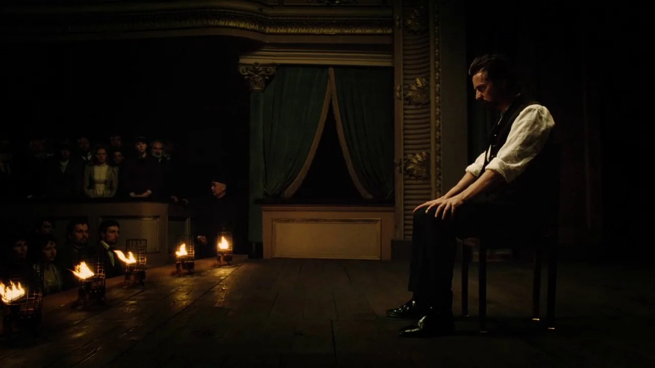 The Illusionist (2006) movie still: medium — A dimly lit wooden stage features an audience seated in shadows behind flaming footlights. A solitary…