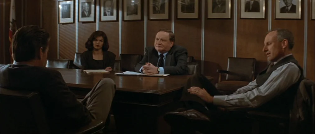 Basic Instinct (1992) movie still: wide — A dark-paneled boardroom with framed photographs on the wall and a central wooden table. Three people face…
