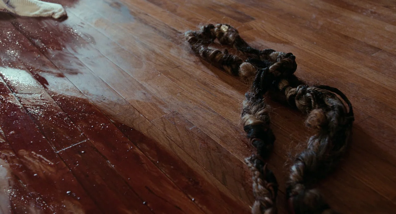 Bones and All (2022) movie still: closeup — A dark-stained wooden floor features a damp patch and a long hair braid. The braid rests directly on the…