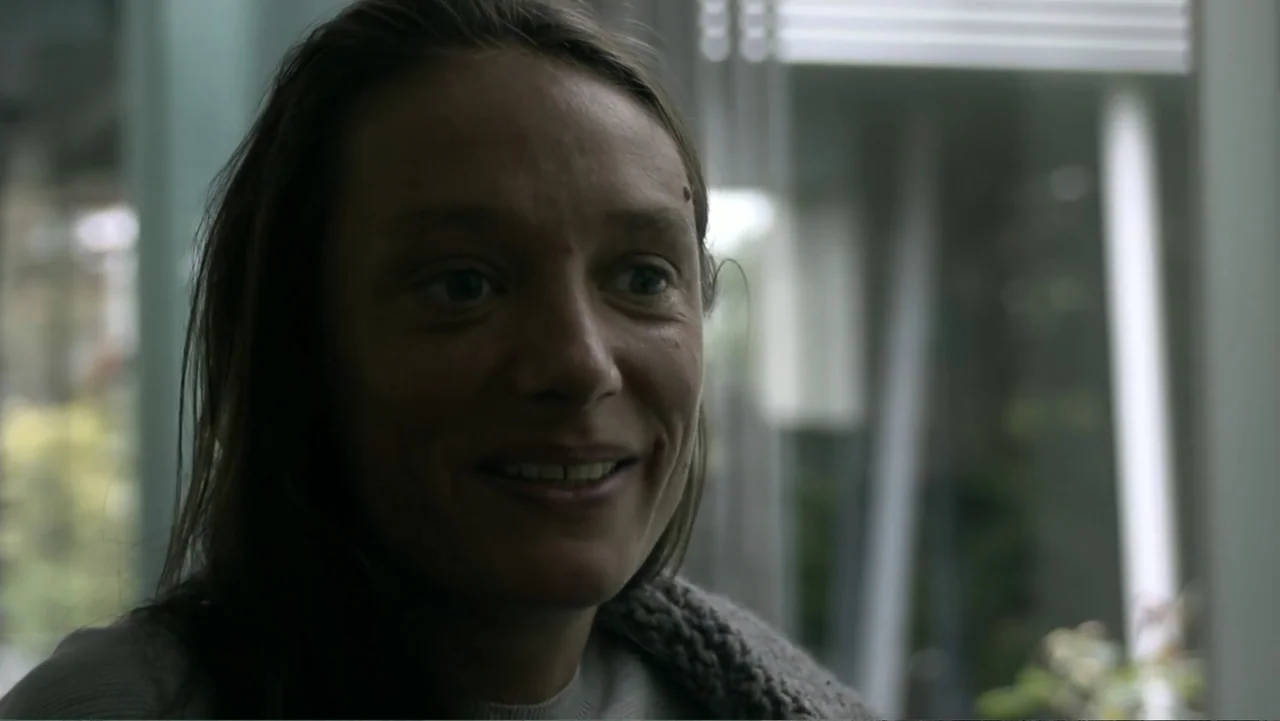 REM (2019) movie still: closeup — A woman's face fills the frame inside a brightly lit modern residential room. She looks off-screen and smiles…