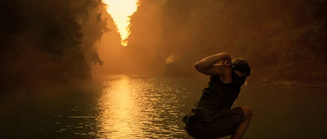 Apocalypse Now (1979) movie still: medium — A wide river flows through a dense jungle under an intense orange fire. A man crouches in the shallow…
