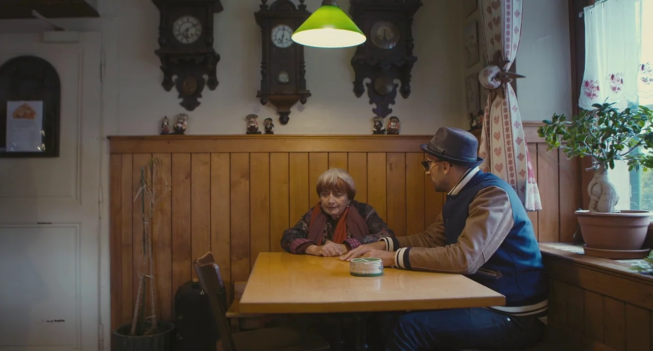 Faces Places (2017) movie still: medium — A rustic wood-paneled room features three antique wall clocks above a square wooden table. An elderly…