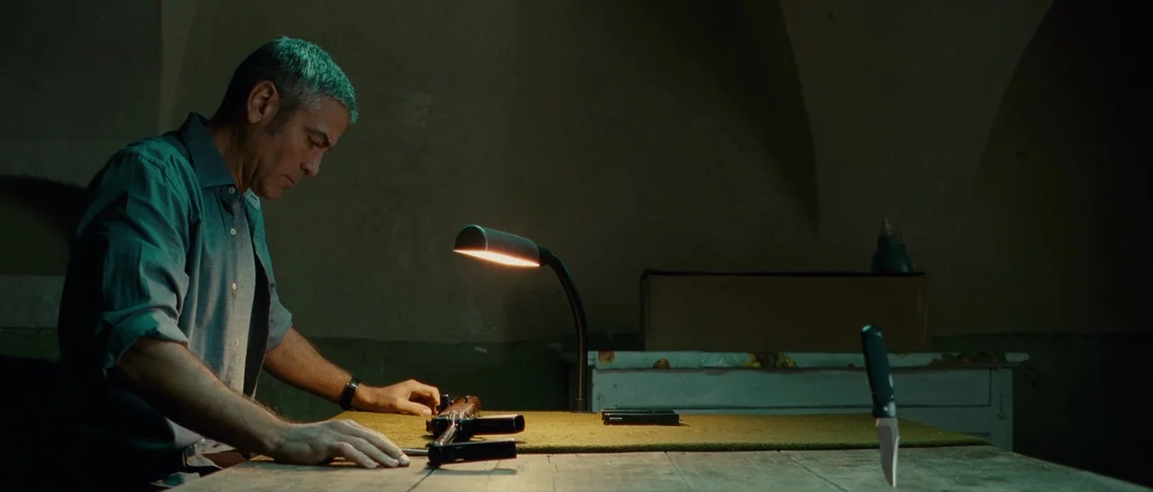 The American (2010) movie still: medium — A dimly lit room with stone walls contains a single table occupied by a man. He looks down at a…