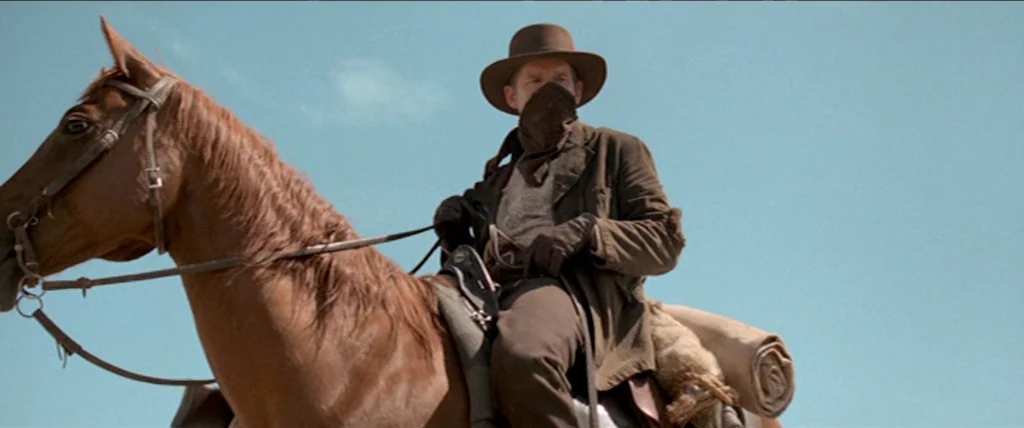 In A Valley of Violence (2016) movie still: medium — A man on horseback moves through a barren area under a clear sky. He wears a wide-brimmed hat…
