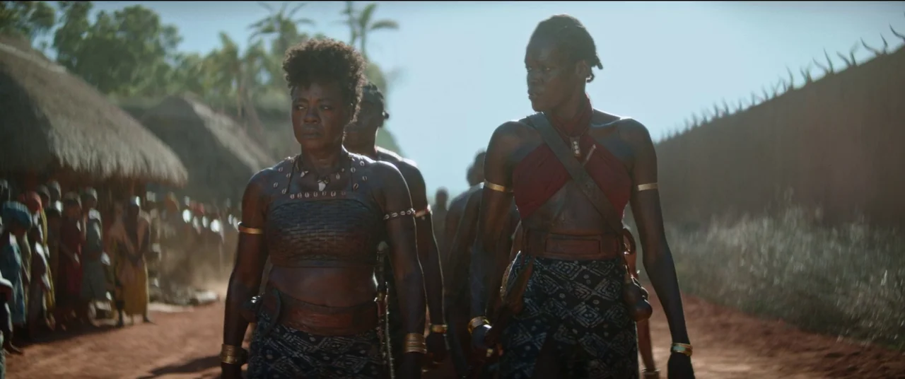 The Woman King (2022) movie still: fullbody — A rural village setting with thatched-roof structures and a high wooden perimeter fence. Two women in…