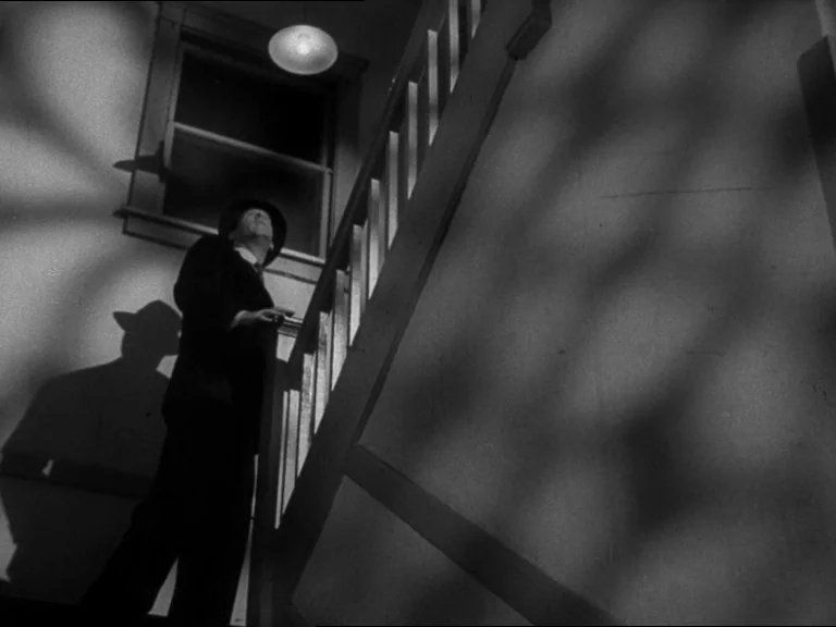 Double Indemnity (1944) movie still: fullbody — An interior staircase with wooden railings and a plain plastered wall behind. A man in a suit looks…