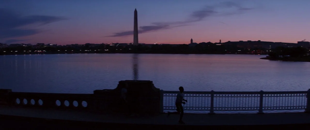 Captain America: The Winter Soldier (2014) movie still: wide — A dark path runs alongside a wide body of water at twilight with a city silhouette. A…