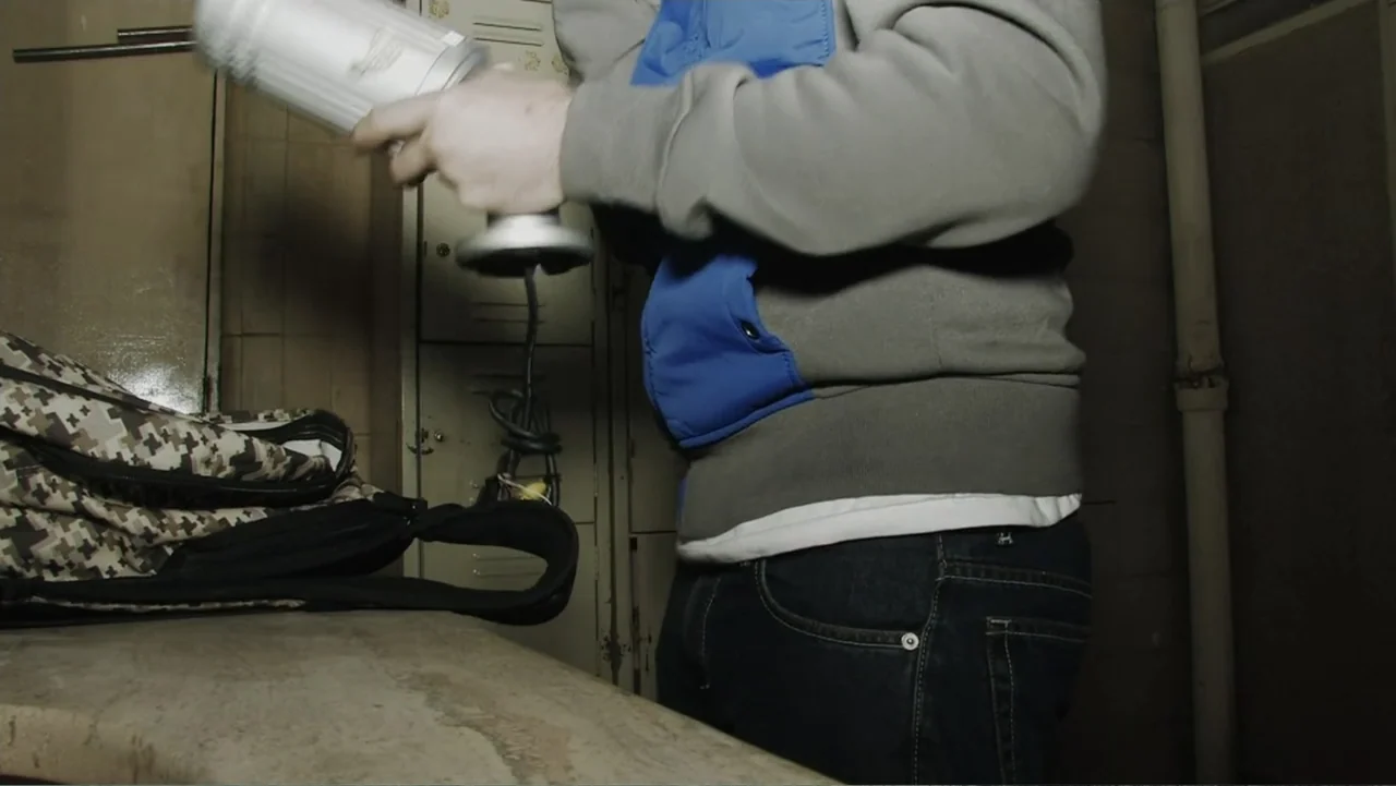 Lost (2010) movie still: medium — A dimly lit locker room features metal lockers and a grey stone counter. A man wearing a grey hoodie handles a…