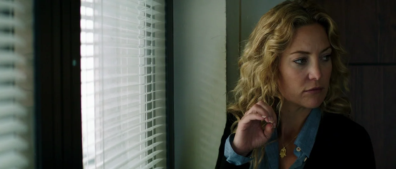 Wish I Was Here (2014) movie still: closeup — An interior office space with a window covered by horizontal plastic blinds. A woman stands near the…