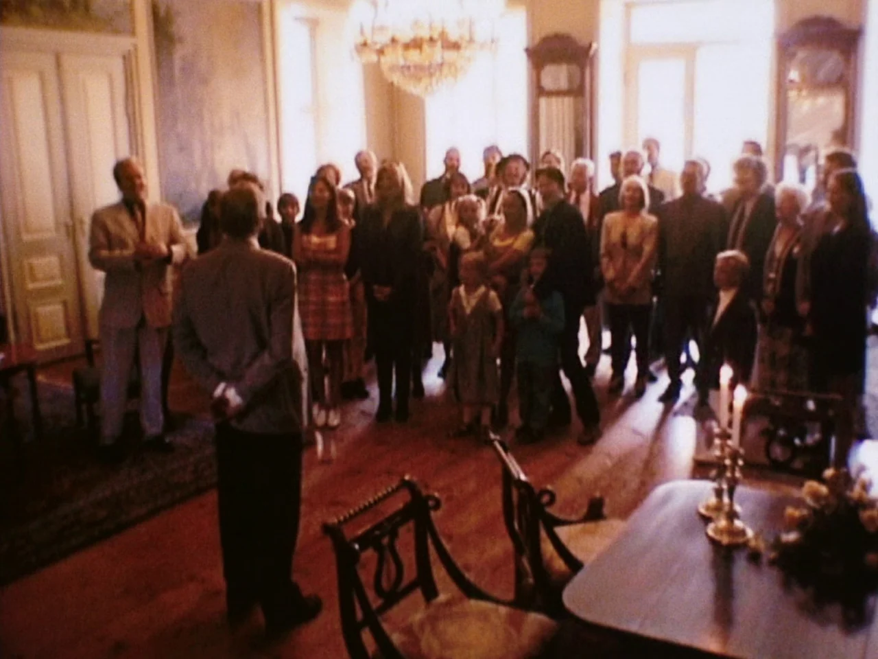 Festen (1998) movie still: wide — A spacious room features polished wooden floors, tall windows, and an ornate crystal chandelier. A large group of…