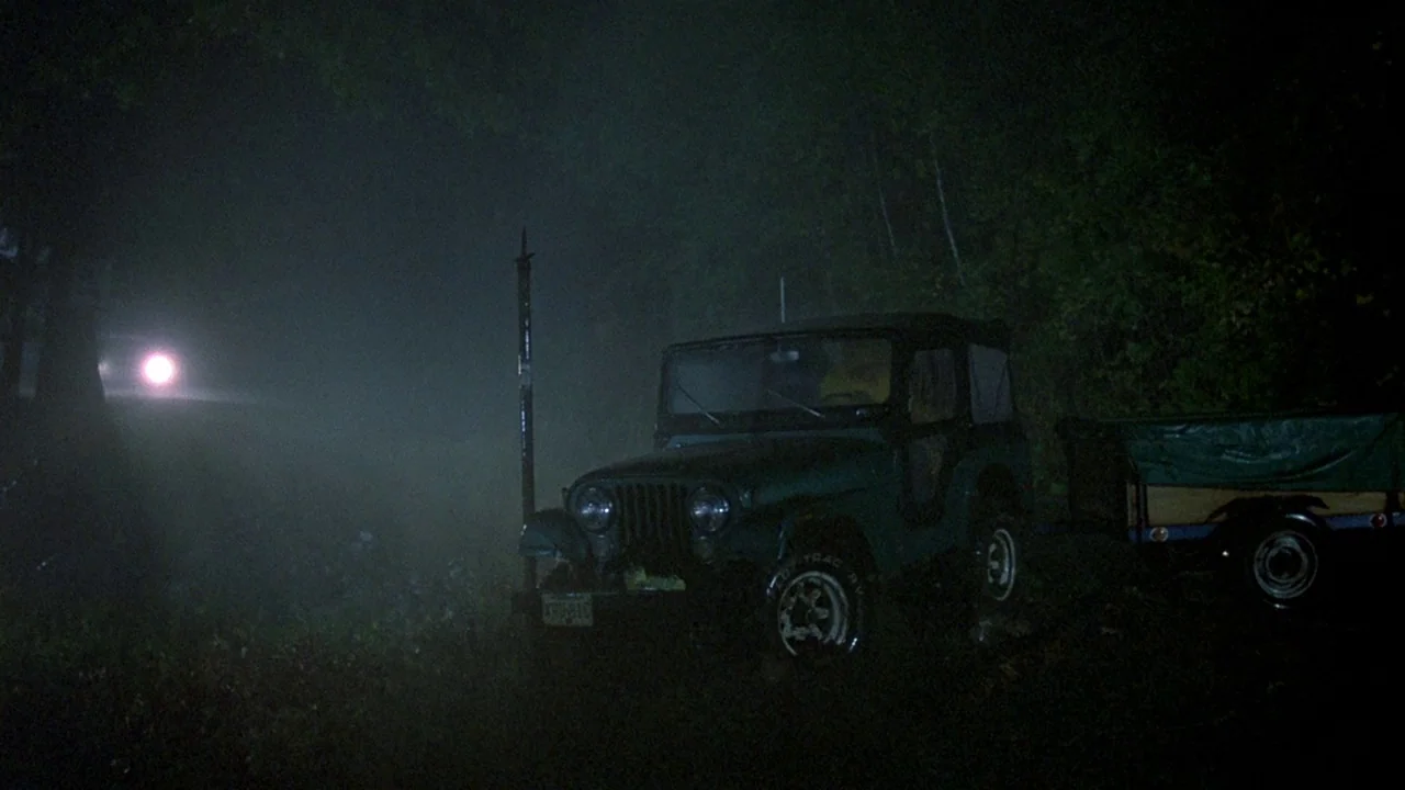 Friday the 13th (1980) movie still: wide — A dark, fog-covered country road features a parked Jeep towing a small trailer. The scene is illuminated…