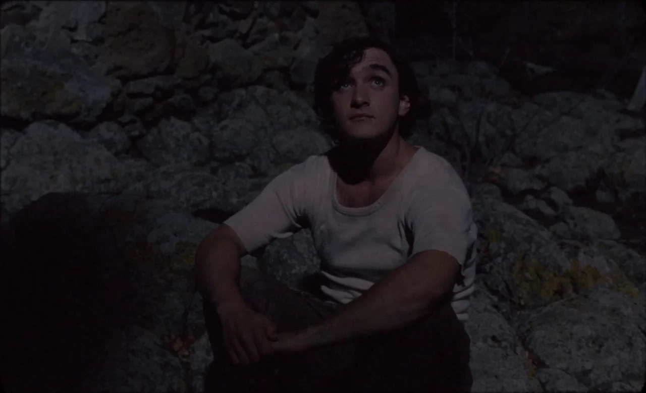 Happy as Lazzaro (2018) movie still: medium — A young man sits on a dark, rugged rock formation at night. He looks upward with a neutral expression.