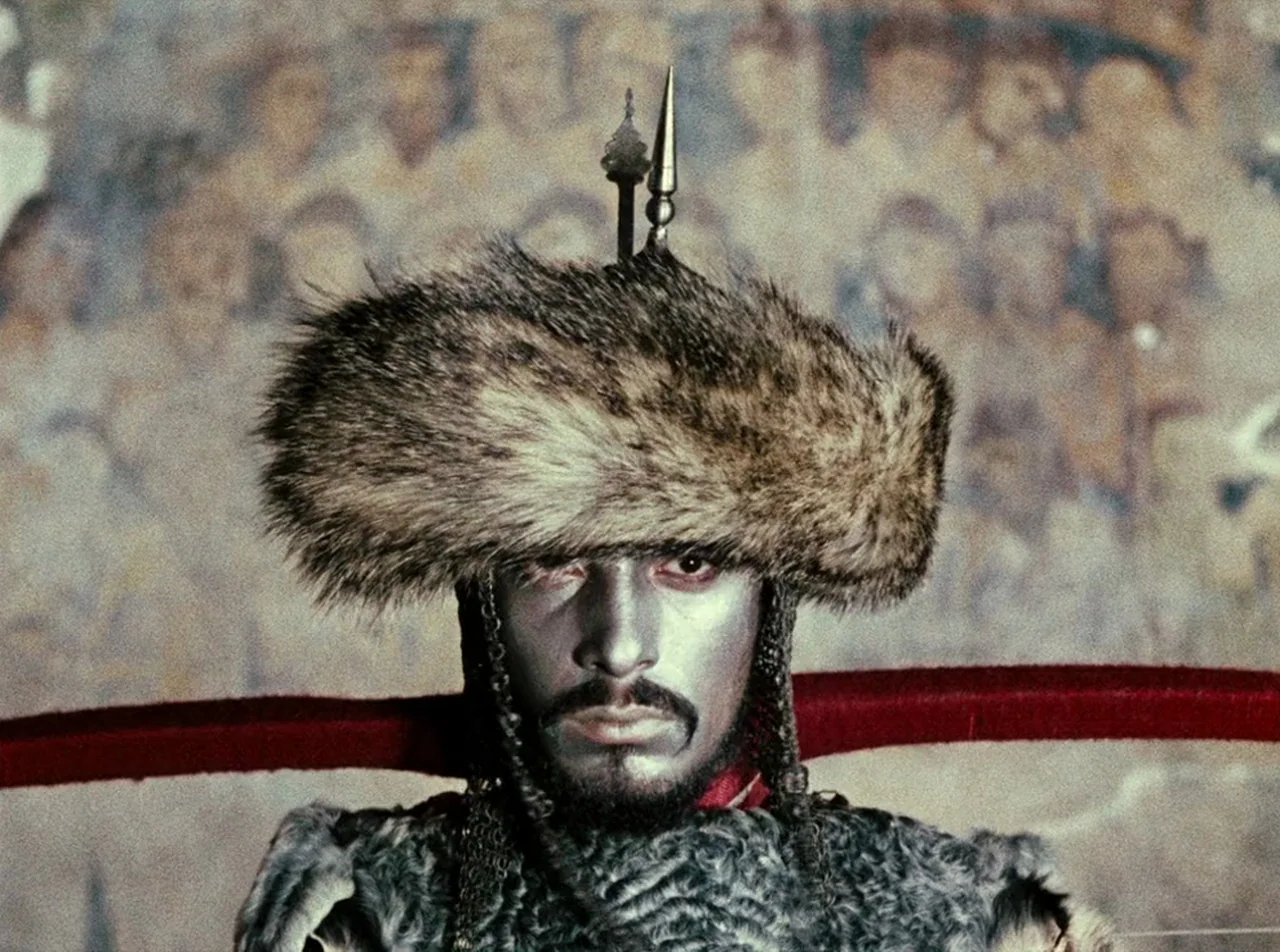 The Colour of Pomegranates (1969) movie still: closeup — A man stands before a blurred mural depicting a crowd of faces. He wears a fur hat and…