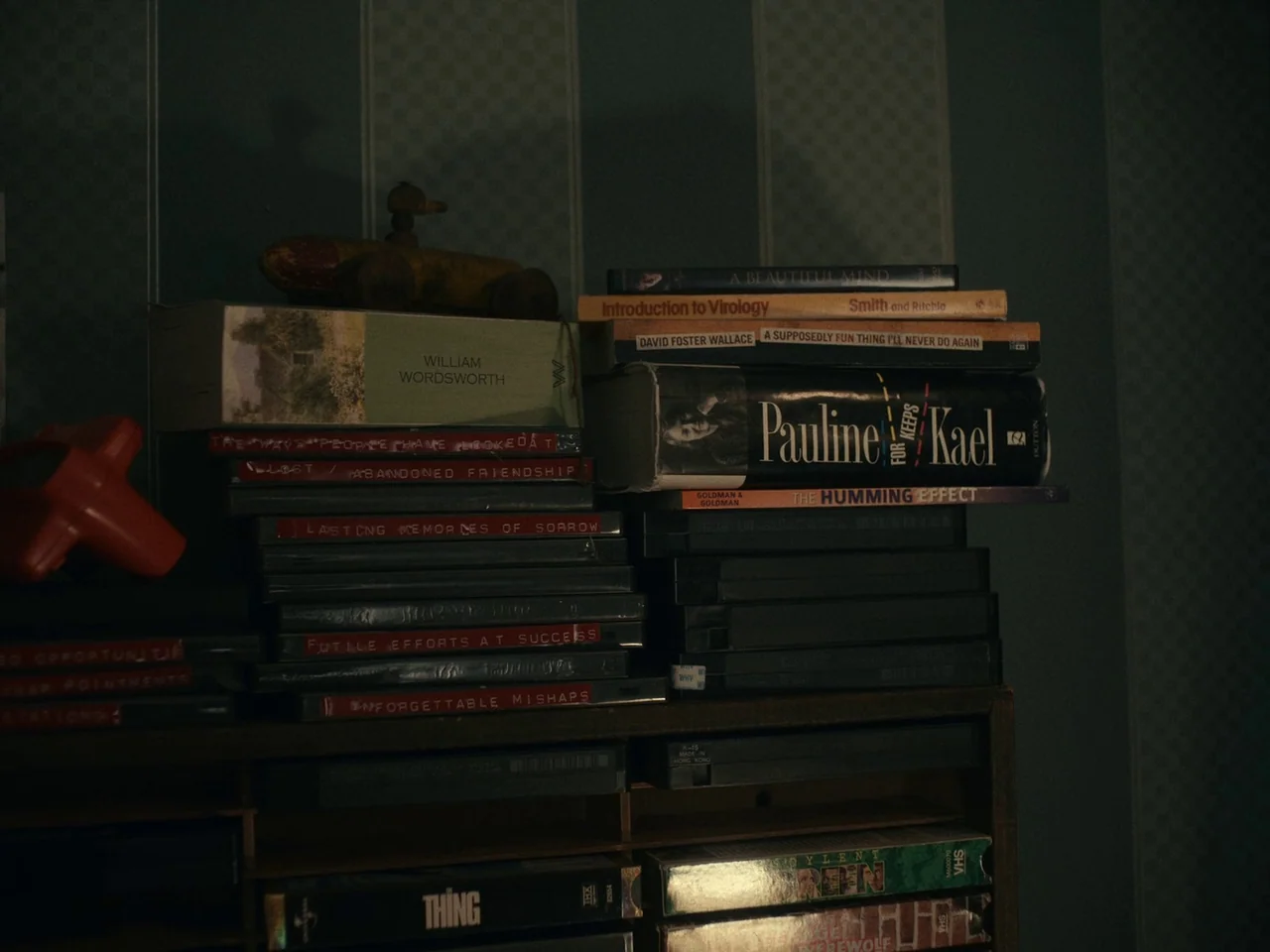 I’m Thinking of Ending Things (2020) movie still: closeup — A dark, wood-framed shelf holds stacks of VHS tapes and hardcover books against…