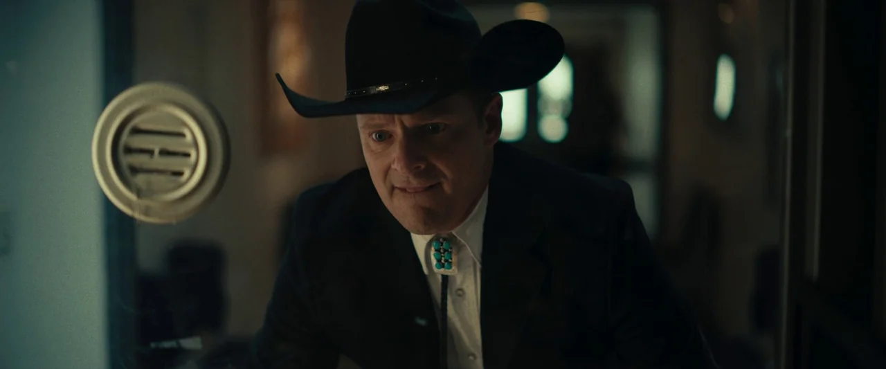 LaRoy, Texas (2023) movie still: medium — A man in a black cowboy hat and suit stands before a glass window. He leans forward with a fixed…