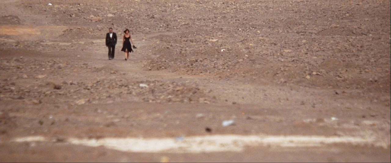 Quantum of Solace (2008) movie still: wide — A vast, empty rocky desert landscape stretches toward a distant horizon under clear skies. A man in a…
