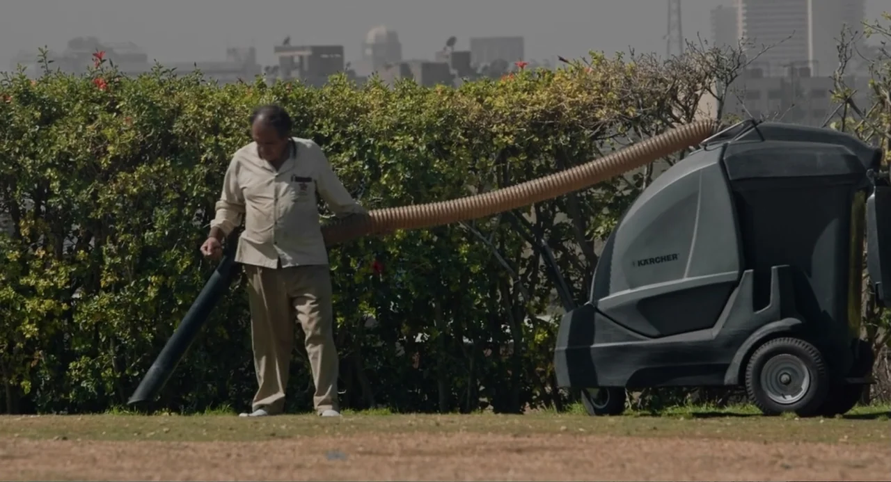 Cairo Jazzman (2017) movie still: fullbody — A sunny outdoor park area features a dense green hedge wall in the background. A man uses a large…