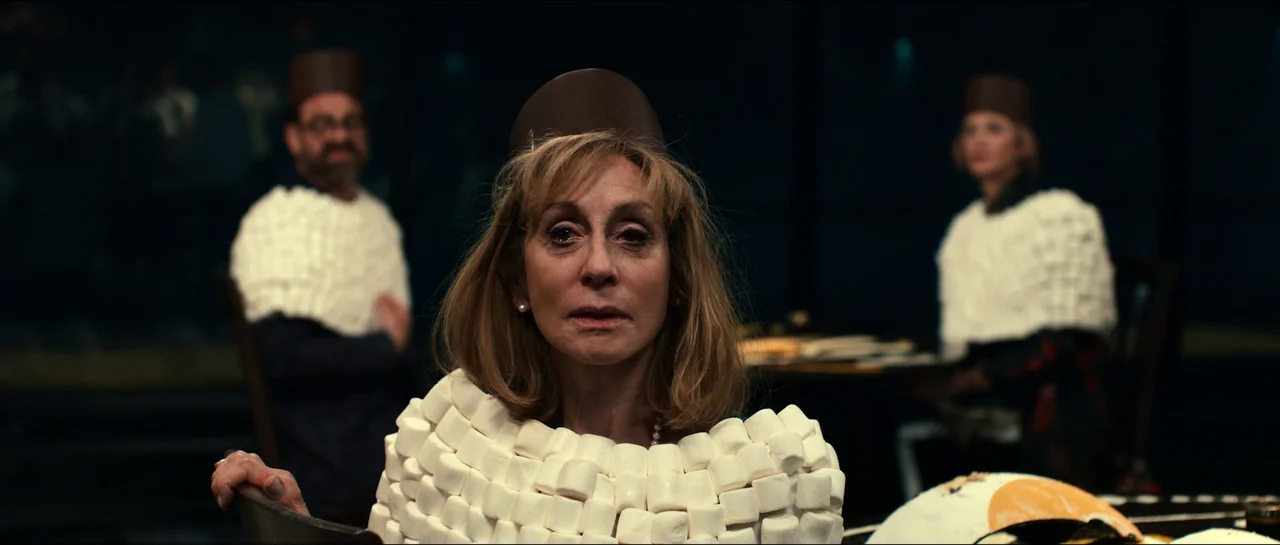 The Menu (2022) movie still: medium — A dimly lit room holds three people seated at a dark table together. Each person wears a custom garment…