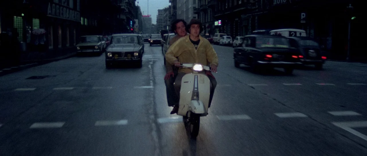The Suspicious Death of a Minor (1975) movie still: fullbody — A wet asphalt city road lined with tall apartment buildings at dusk. Two men ride a…