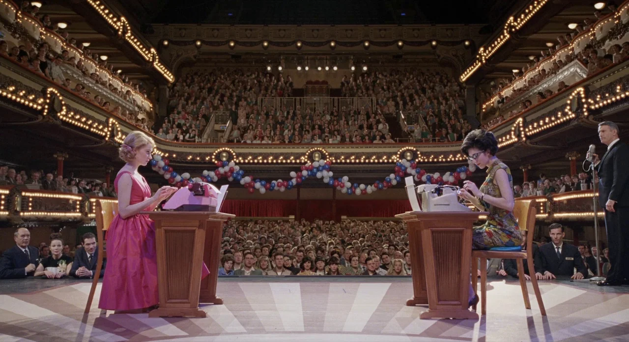 Populaire (2012) movie still: wide — A crowded vintage theater features a woman at each of two desks facing typewriters. The two women compete in a…
