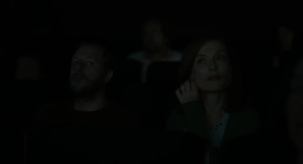 Things to Come (2016) movie still: medium — A dark, dimly lit movie theater interior with rows of padded seats and occupants. A man and a woman sit…