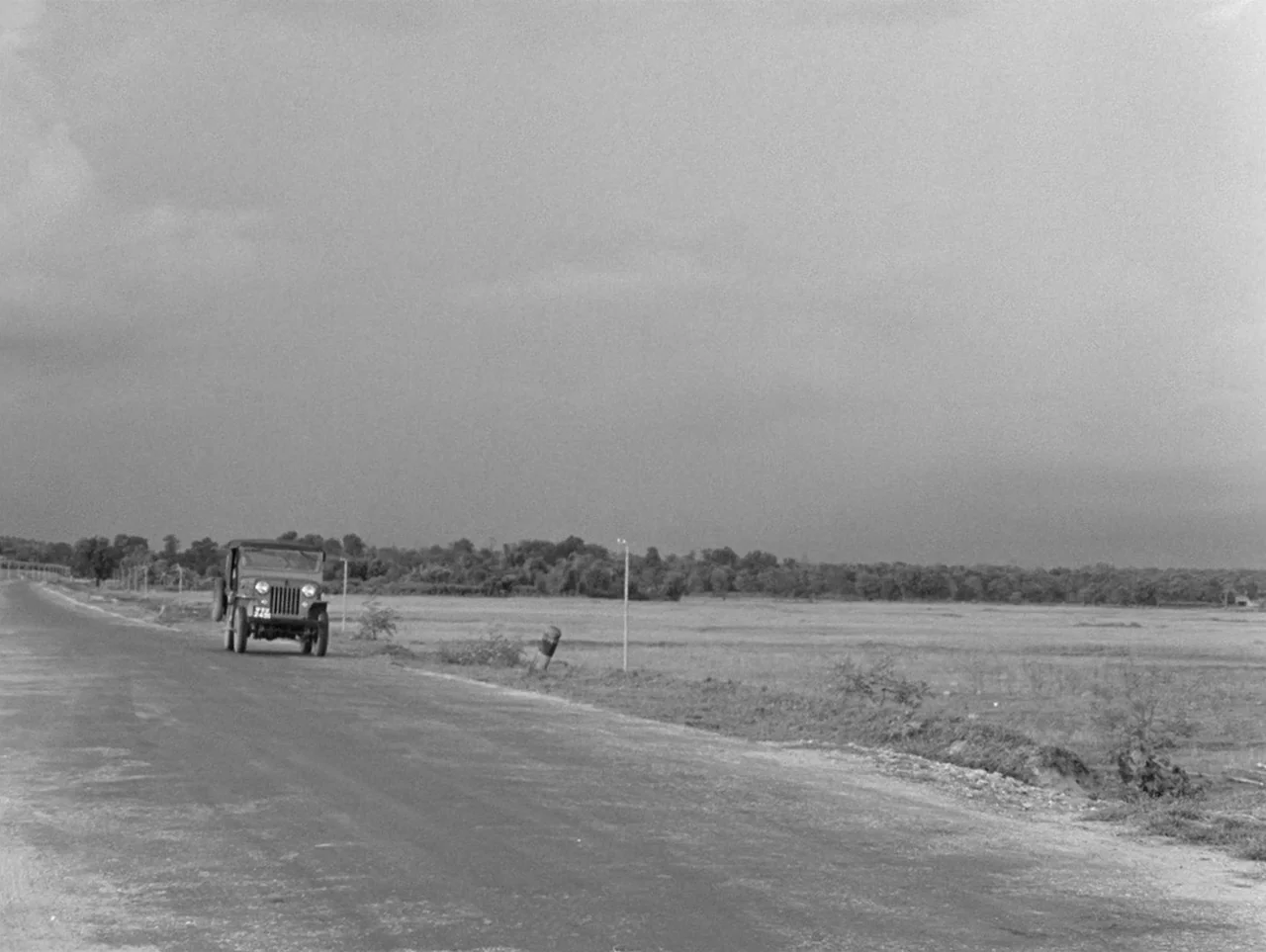 The Coward (1965) movie still: wide — An open rural landscape features a paved road flanked by flat, dry grassy fields. A small vintage jeep travels…