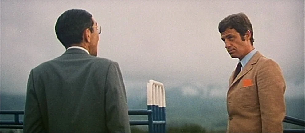 Mississippi Mermaid (1969) movie still: medium — Two men talk on an outdoor balcony overlooking a hazy, grey landscape. A blue metal railing stands…