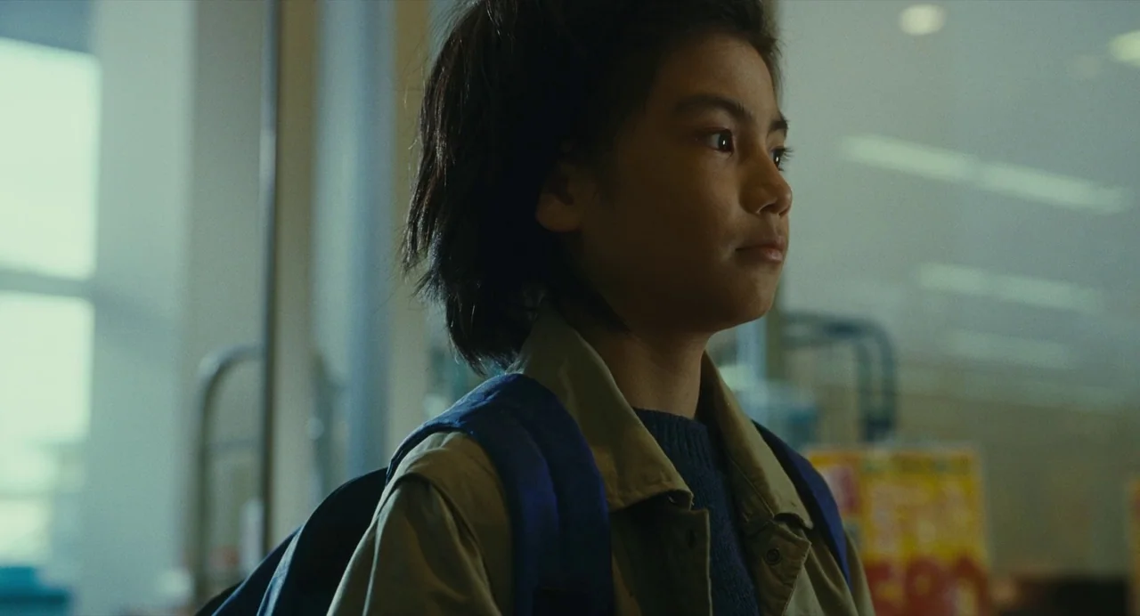 Shoplifters (2018) movie still: medium — A well-lit commercial space features glass shelving and signs in the blurred background. A young child with…