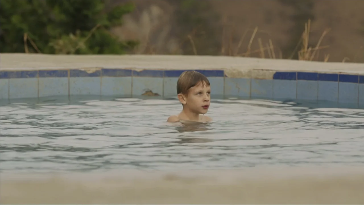 Boy (2010) movie still: medium — A worn outdoor swimming pool with blue tiles and a cracked concrete deck. A young boy stands in the water, looking…