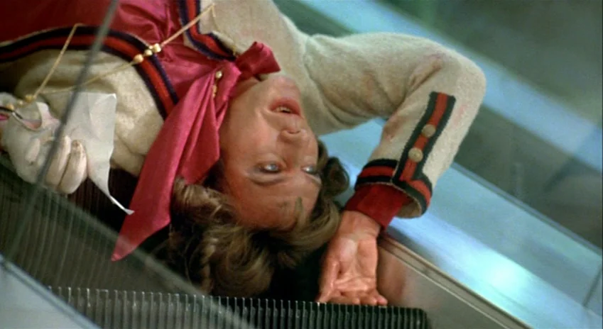 The Silent Partner (1978) movie still: medium — A person lies across the metal steps of an escalator in a public space. The subject stares upward…