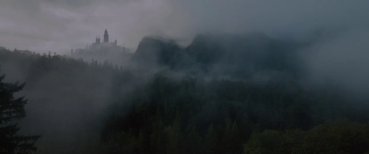 Ophelia (2018) movie still: establishing — A silhouette of a castle sits atop a misty mountain ridge overlooking a forest. Dense fog partially…