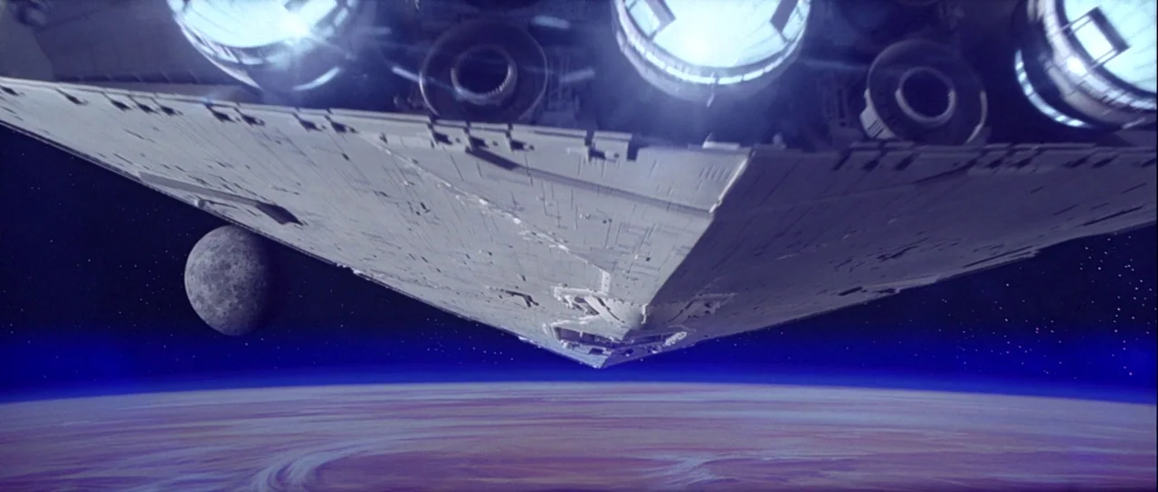 Star Wars (1977) movie still: establishing — A massive, wedge-shaped star destroyer hangs in the dark void of space above a planet. The ship's…