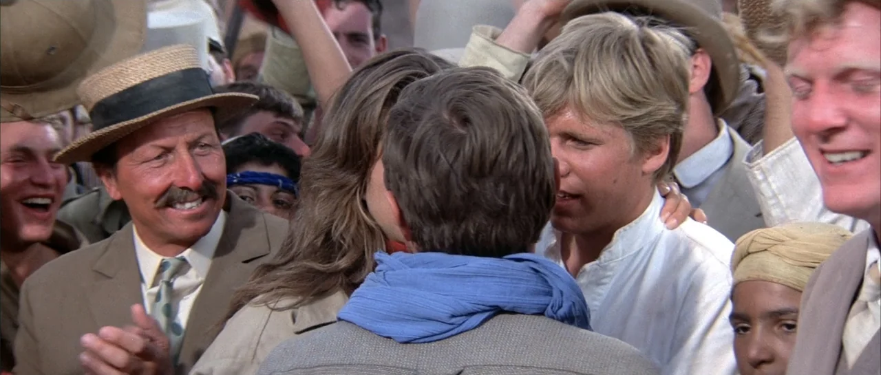 Sahara (1995) movie still: medium — A diverse crowd of people in 1930s style clothing gathers outdoors during the day. A couple stands in the center…