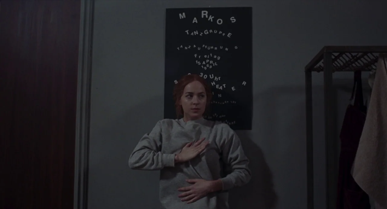 Suspiria (2018) movie still: medium — A dimly lit room features a grey wall with a black dance advertisement poster. A woman stands in front of the…