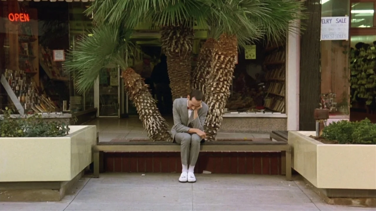 Pee-Wee’s Big Adventure (1985) movie still: fullbody — A shopping plaza features a tiled walkway with large planter boxes and palm trees. A man in a…