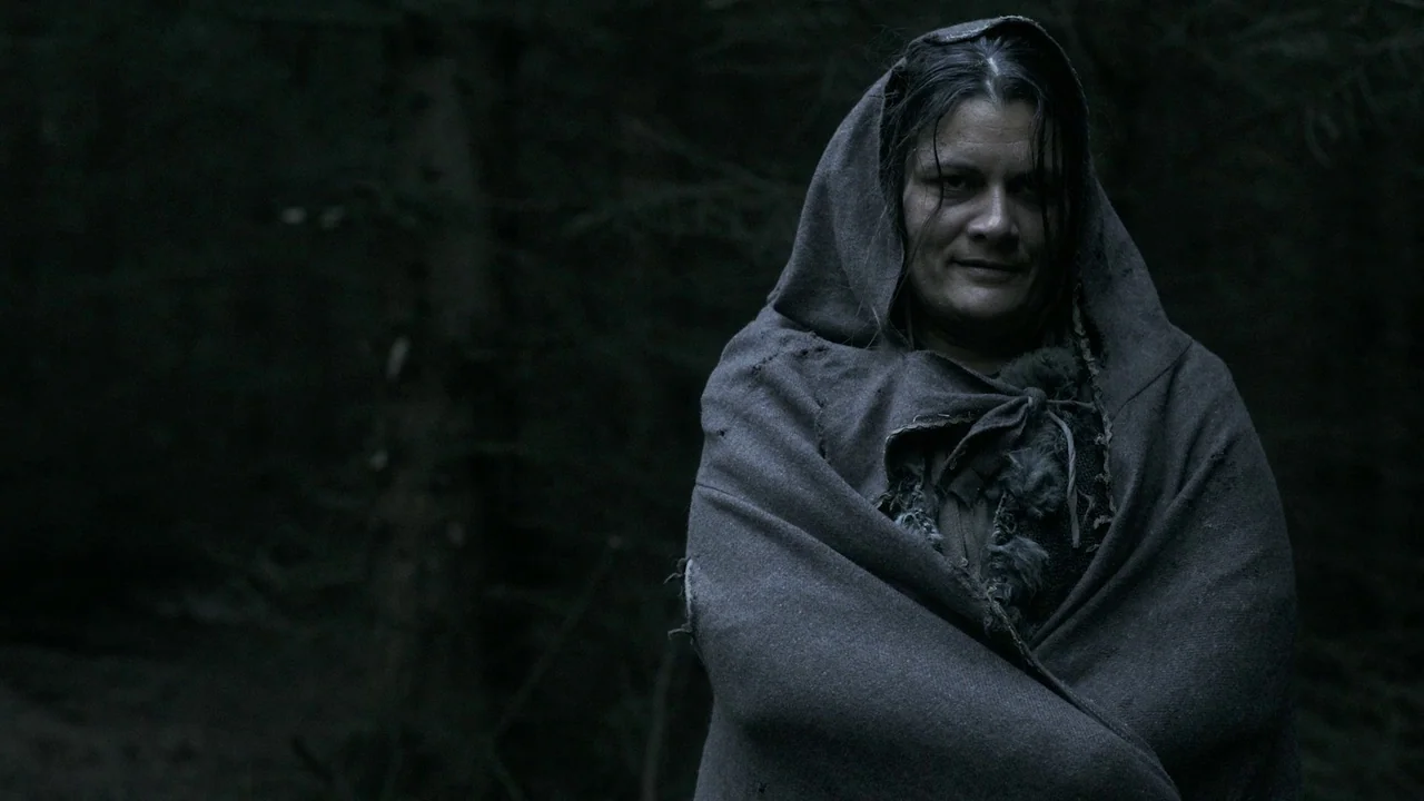 Helll Hole (2024) movie still: medium — A woman stands in a dark, out-of-focus woodland area at night. She wears a worn cloak while facing the camera.