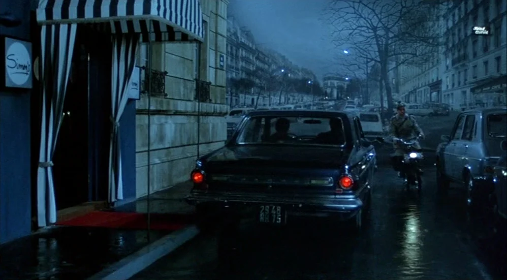 Un Flic (1972) movie still: wide — A Parisian street at night features a car parked in front of a building. A motorcyclist travels down the road…