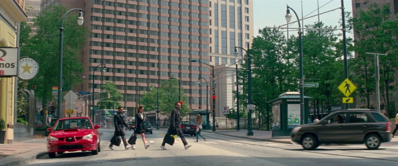 Baby Driver (2017) movie still: wide — An urban intersection with high-rise buildings and green trees lining the street. Three individuals in dark…