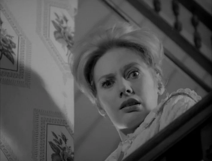 Carnival of Souls (1962) movie still: closeup — A woman stares down from a dark, ornate wooden staircase with floral wallpaper nearby. The subject…